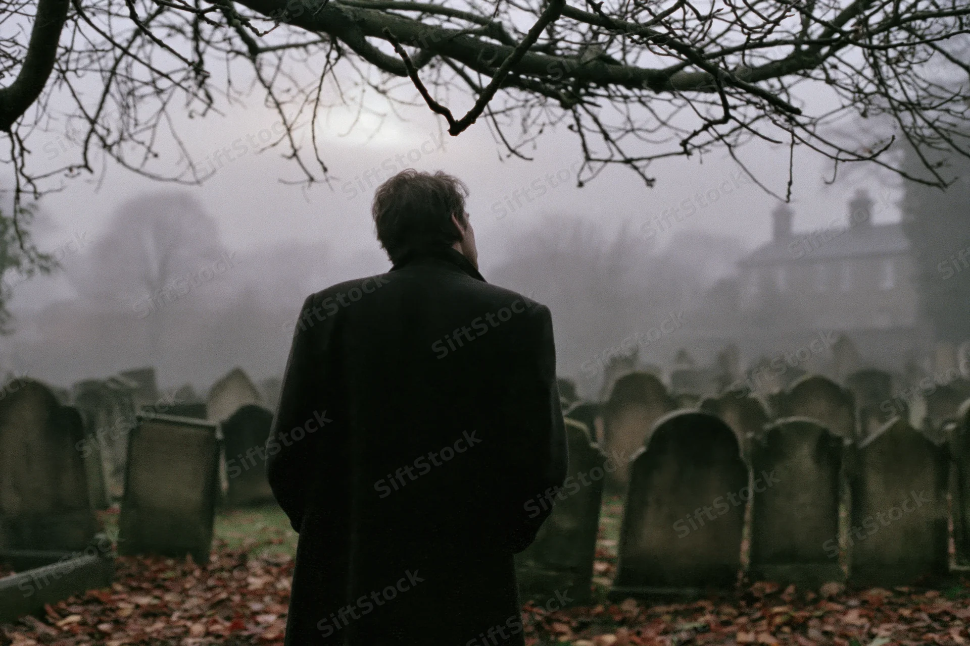 Man in dark overcoat standing with back to camera in foggy cemetery among gravestones