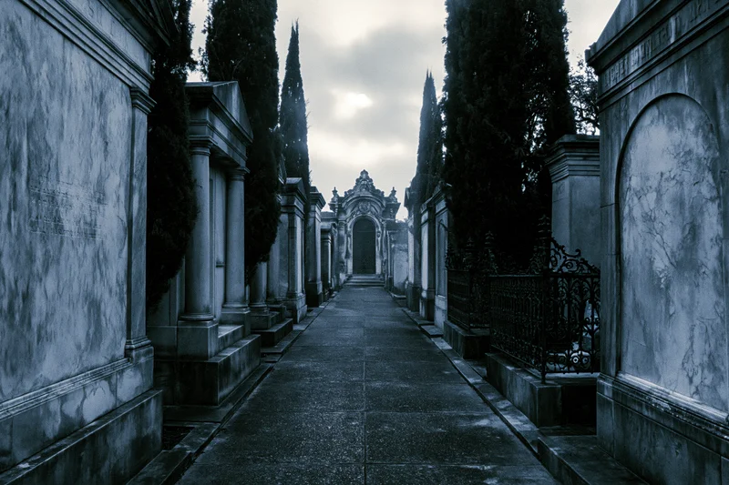 Narrow stone path through cemetery flanked by old marble mausoleums under overcast sky