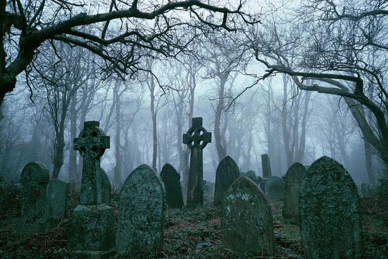 Weathered gravestones and celtic crosses in a foggy cemetery surrounded by bare trees