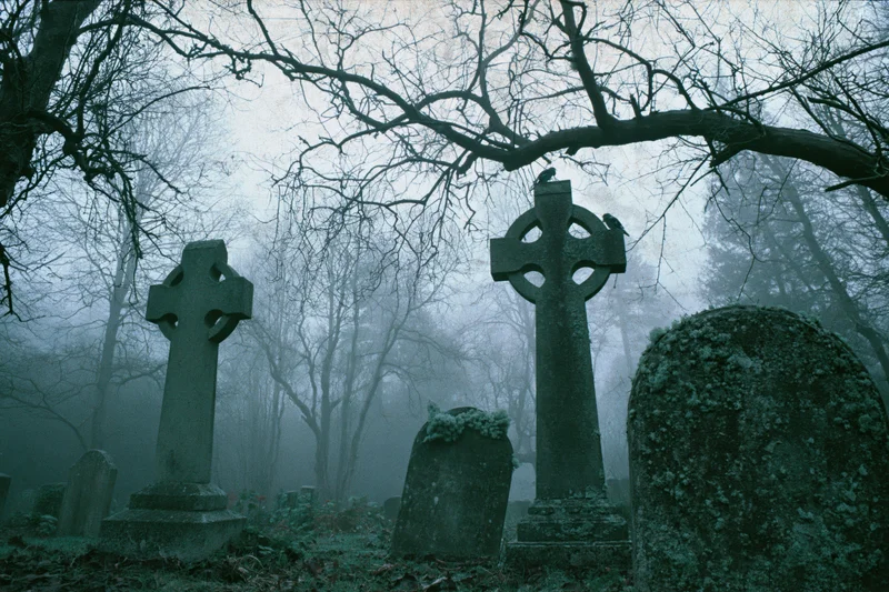 Weathered celtic crosses and gravestones in a foggy cemetery surrounded by bare trees