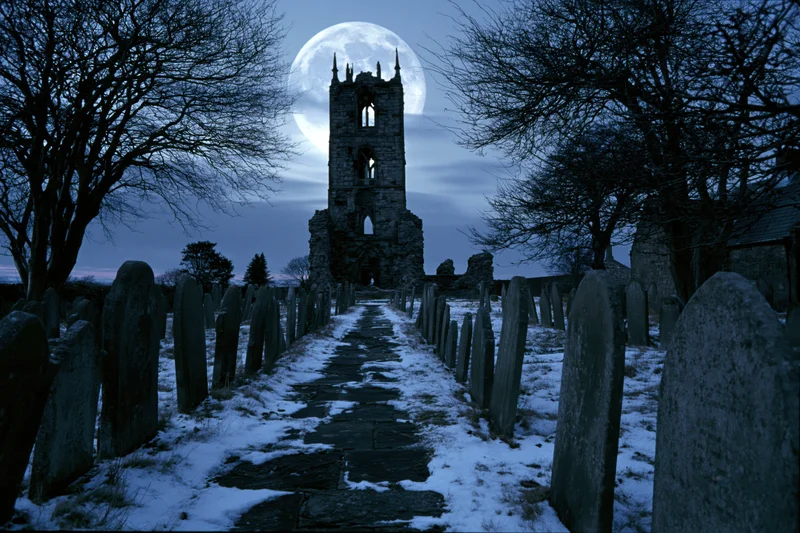 Snow-covered pathway lined with aged headstones leading to a ruined stone tower under a full moon at night
