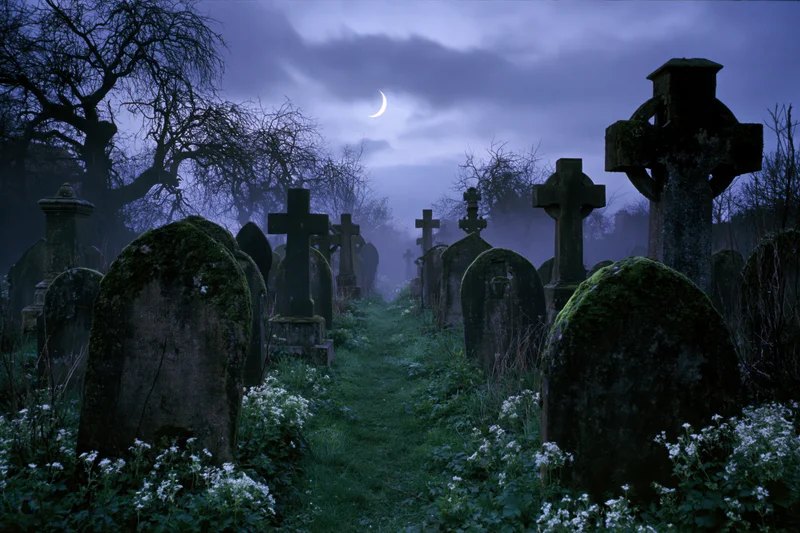 Moss-covered headstones and stone crosses in a foggy graveyard at night under a crescent moon