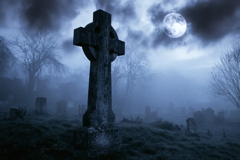 Old stone cross gravestone standing in a foggy cemetery at night under a full moon