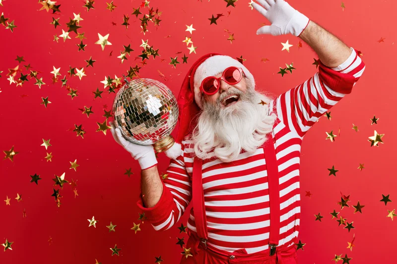 Older man dressed as santa claus in striped shirt and suspenders holding disco ball with confetti falling around him