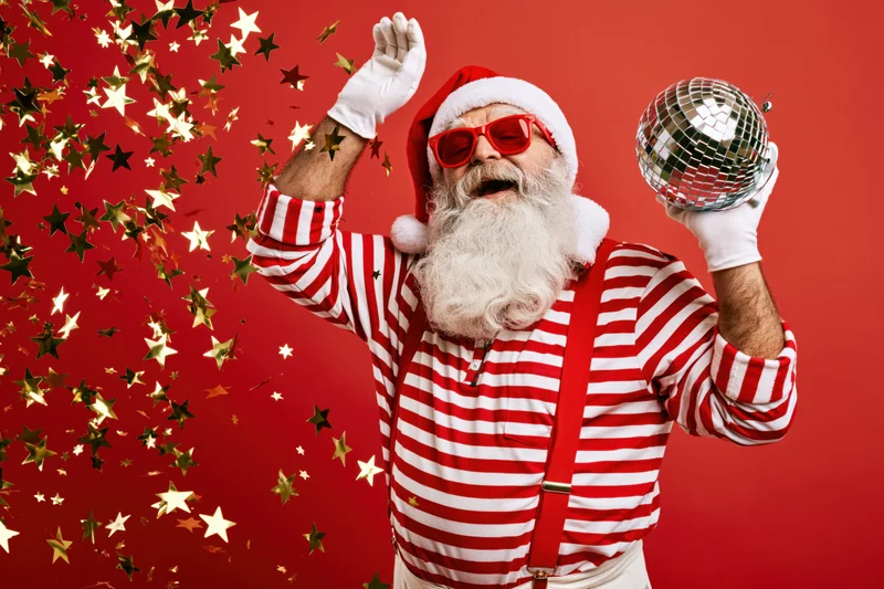 Older man dressed as santa claus in striped shirt and sunglasses dancing with disco ball and confetti against red background