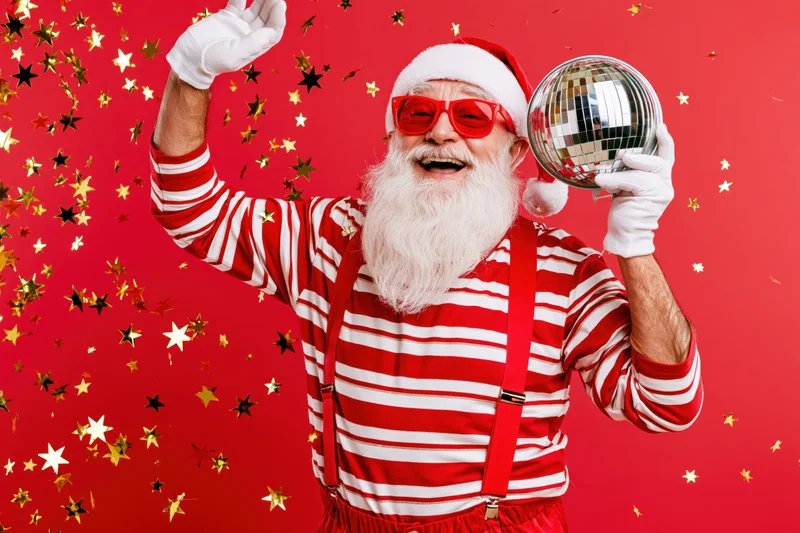 Older man dressed as santa claus in red and white striped shirt with suspenders, holding a disco mirrorball and waving, surrounded by falling star confetti against a red background