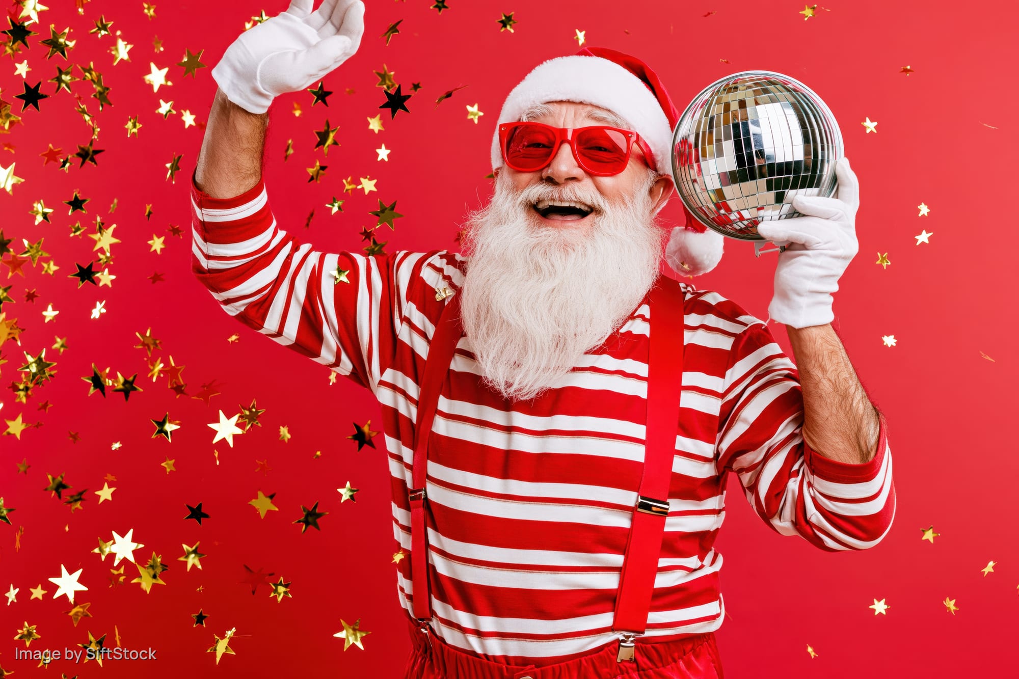 Older man dressed as santa claus in red and white striped shirt with suspenders, holding a disco mirrorball and waving, surrounded by falling star confetti against a red background