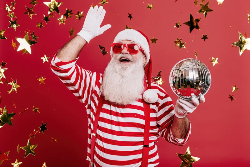 Man dressed as santa claus waving and holding a disco ball against a red background with falling stars