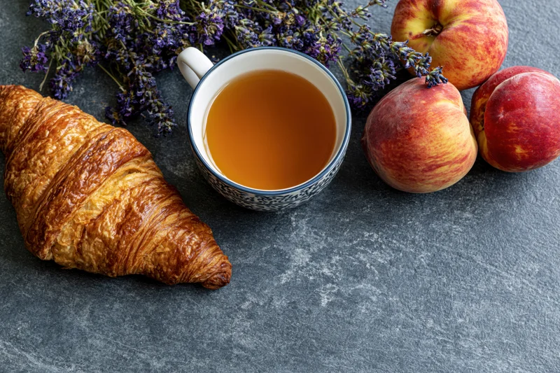 Golden croissant, patterned ceramic cup of tea, peaches and lavender arranged on slate surface for breakfast flat lay