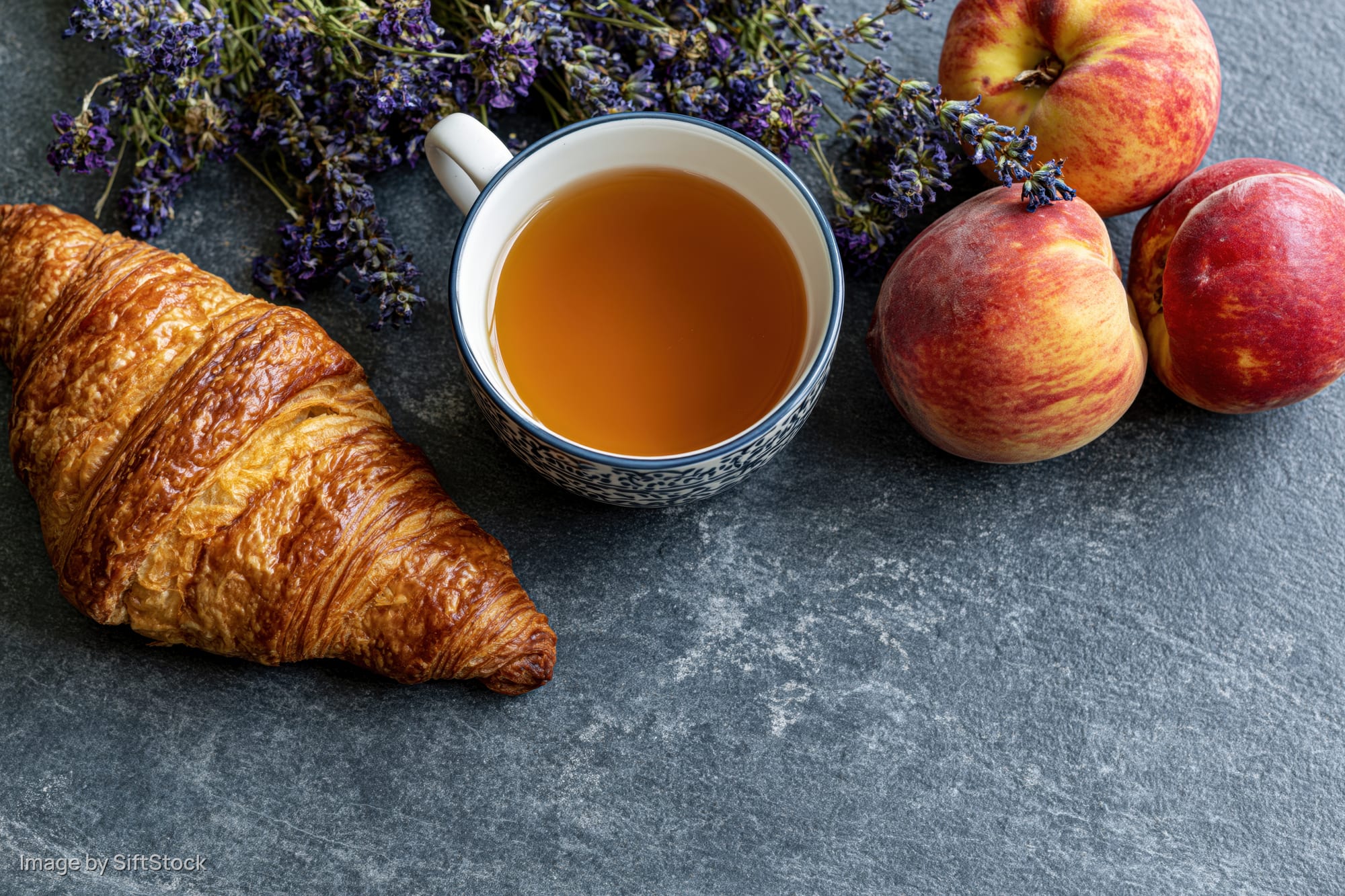 Golden croissant, patterned ceramic cup of tea, peaches and lavender arranged on slate surface for breakfast flat lay