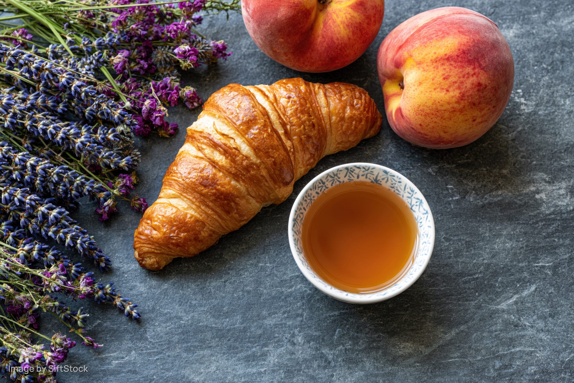 Golden croissant with tea, peaches and lavender arranged on a stone surface for breakfast flat lay