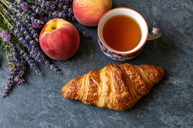 Golden croissant with tea, peaches and lavender arranged on dark stone surface in overhead flat lay