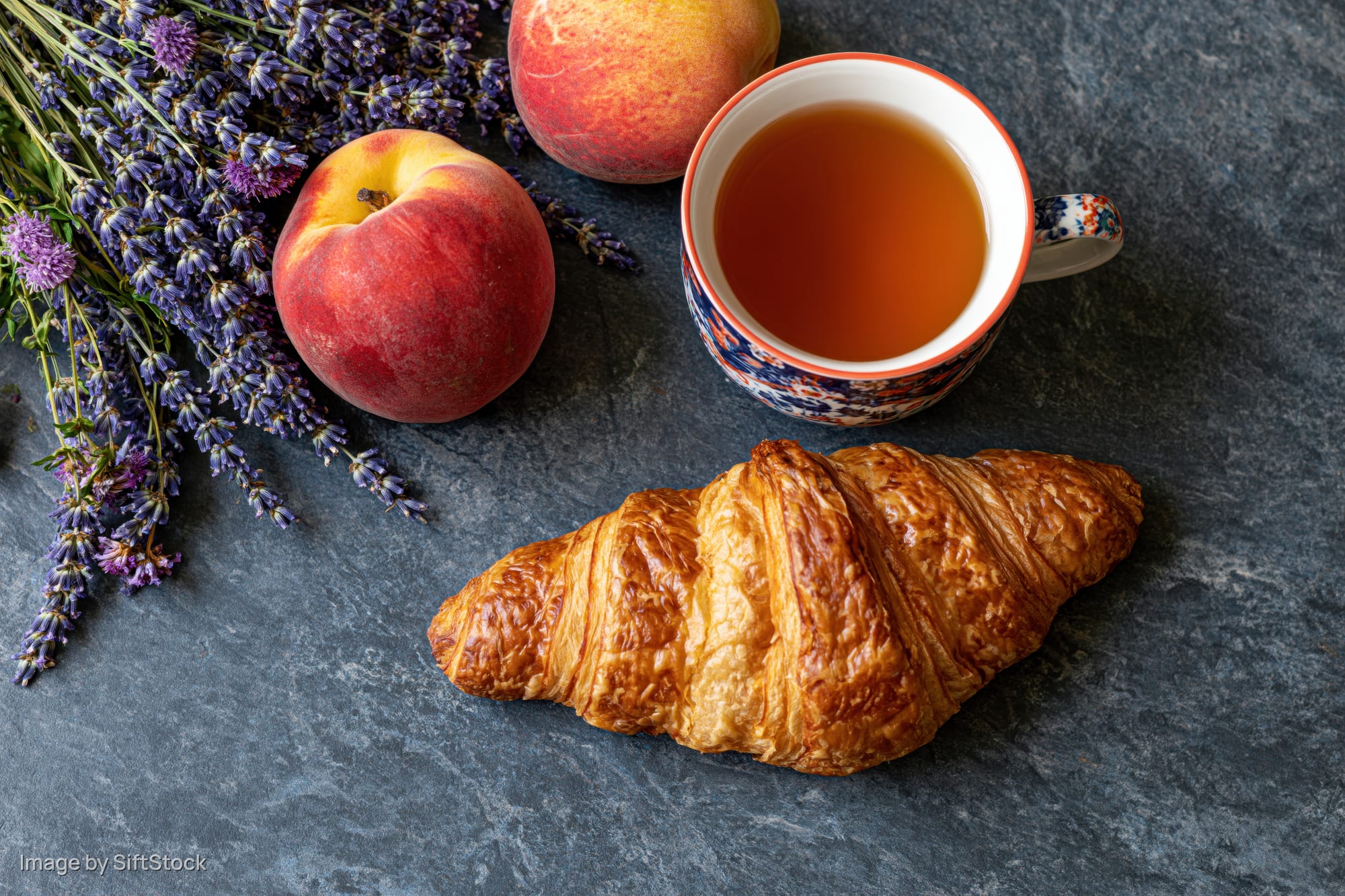 Golden croissant with tea, peaches and lavender arranged on dark stone surface in overhead flat lay
