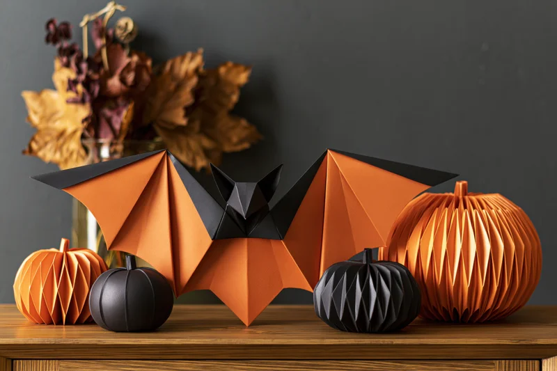 Origami bat and pumpkins arranged on a wooden shelf with dried autumn leaves in a vase behind