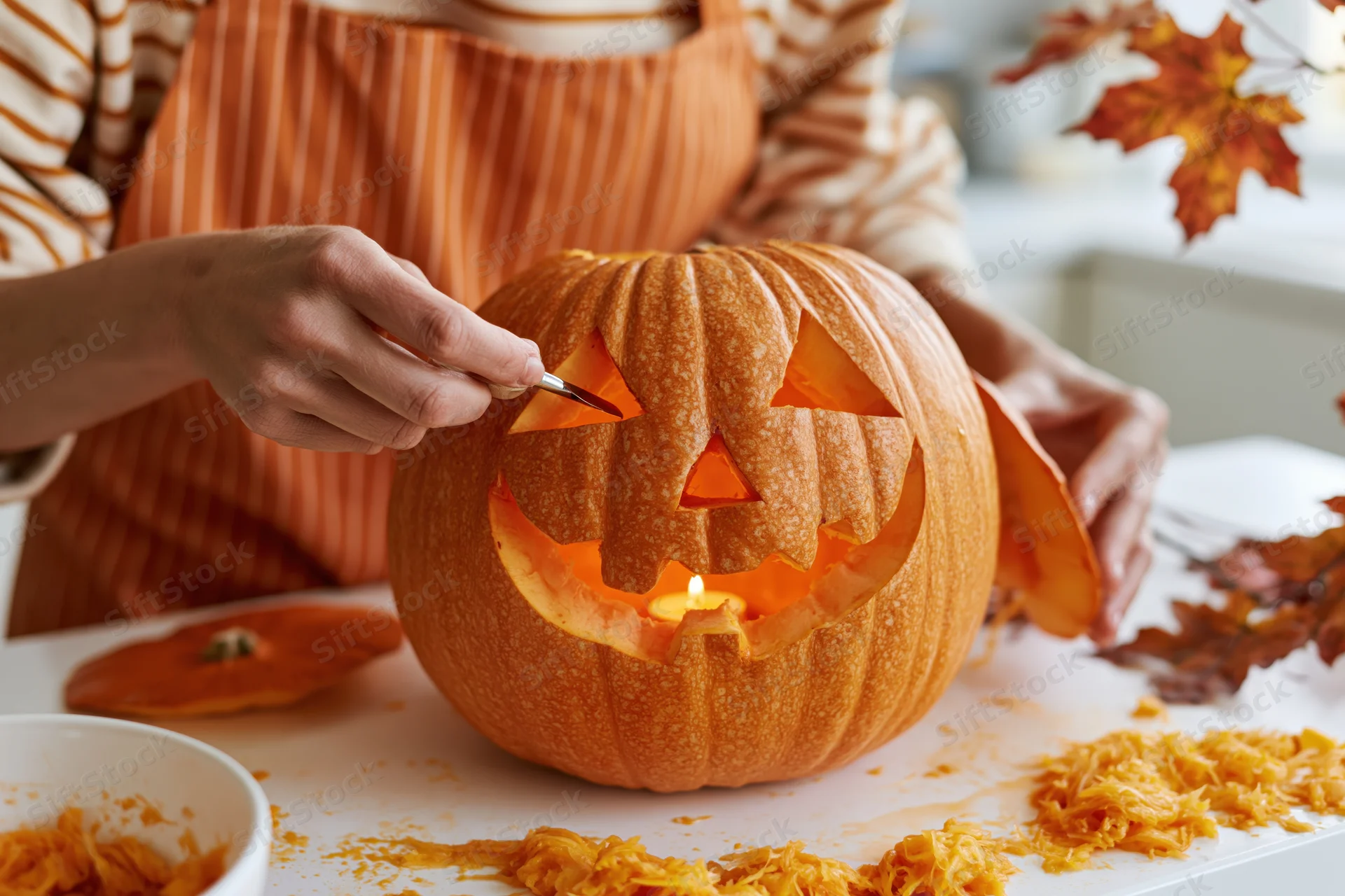 Woman carving a jack-o-lantern with a small knife on a white kitchen table surrounded by autumn leaves and pumpkin pulp