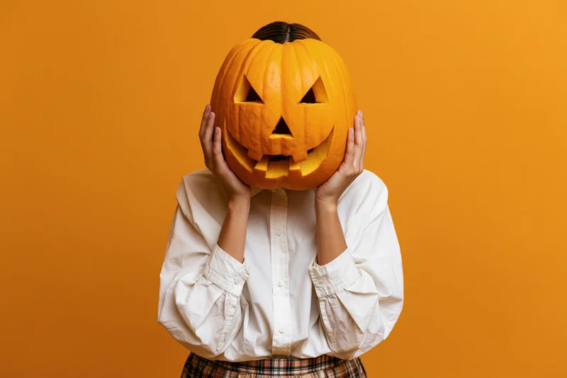 Woman holding carved pumpkin over face against orange backdrop