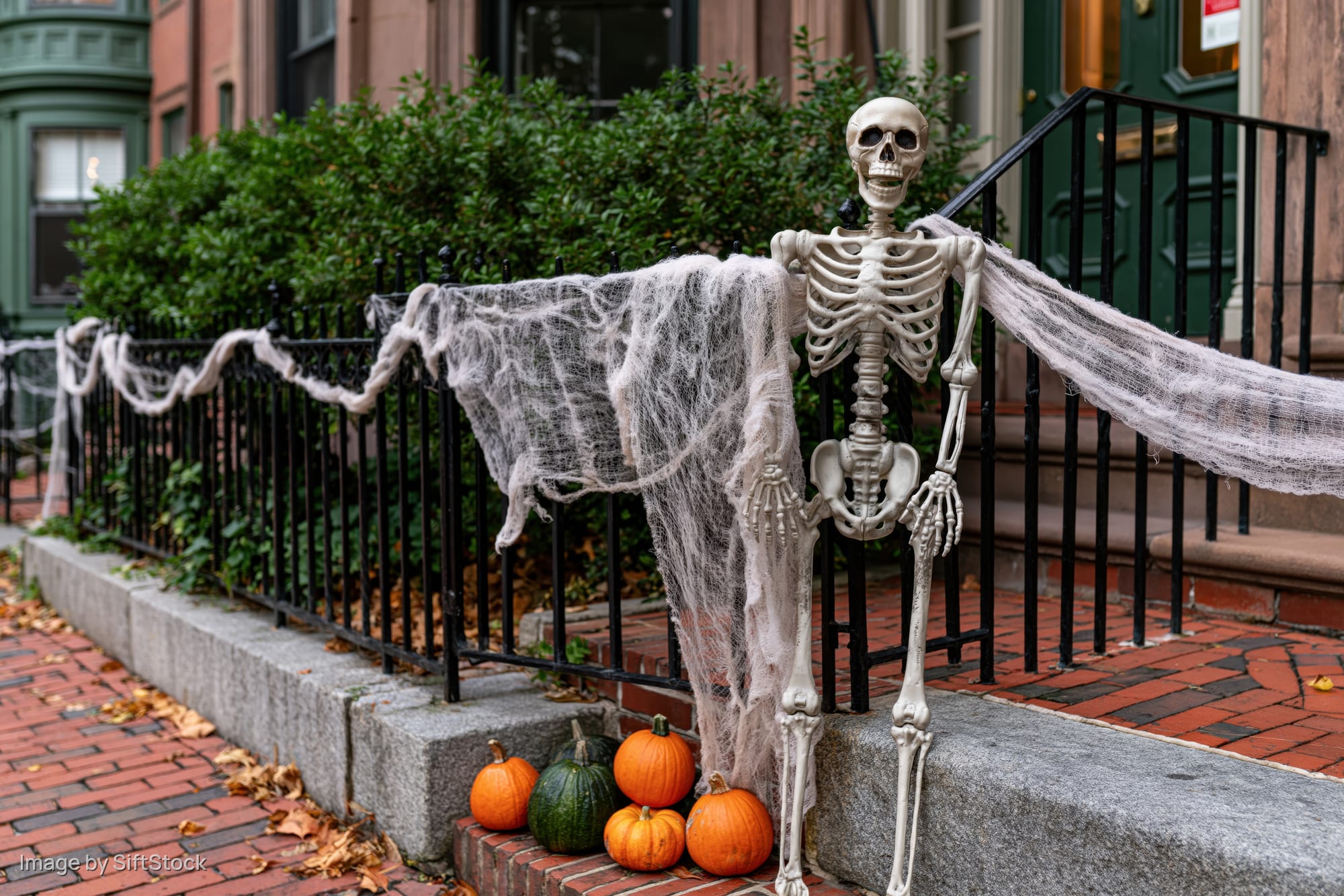 Plastic skeleton draped in fake cobwebs on front porch railing with pumpkins and autumn leaves on brick steps