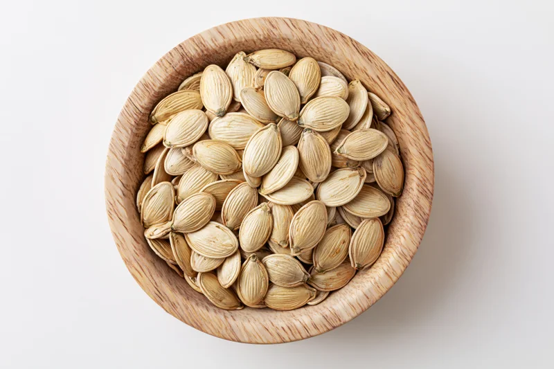 Raw pumpkin seeds filling a small round wooden bowl on white surface