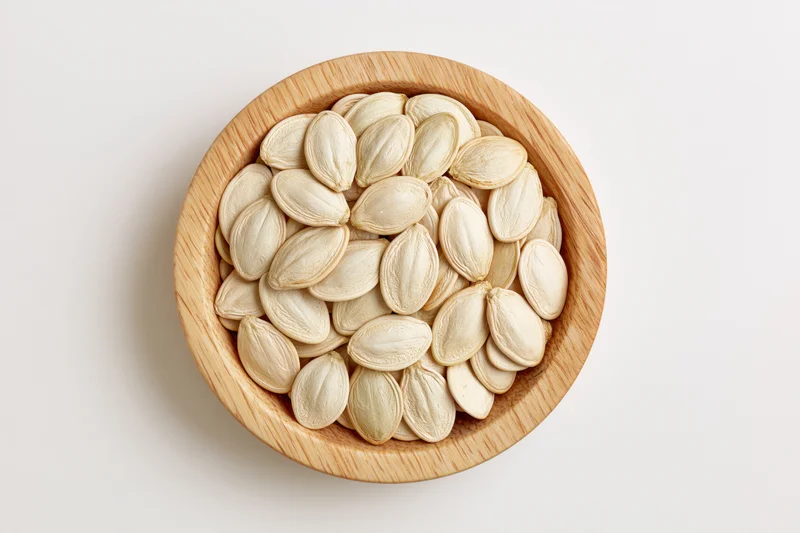 Raw pumpkin seeds filling a small round wooden bowl on a white surface