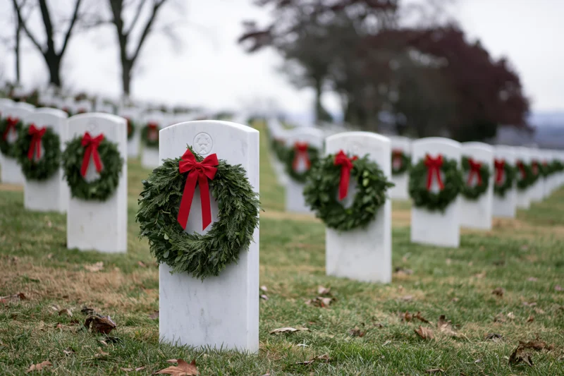 Rows of white marble headstones in a military cemetery adorned with green wreaths and red bows on grassy ground