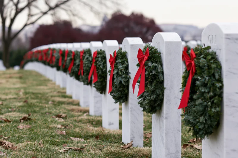 Rows of white marble headstones in a military cemetery adorned with evergreen wreaths and red ribbons during winter