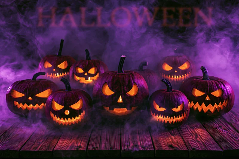 Seven carved jack-o-lanterns glowing with orange light on a wooden table in purple smoke