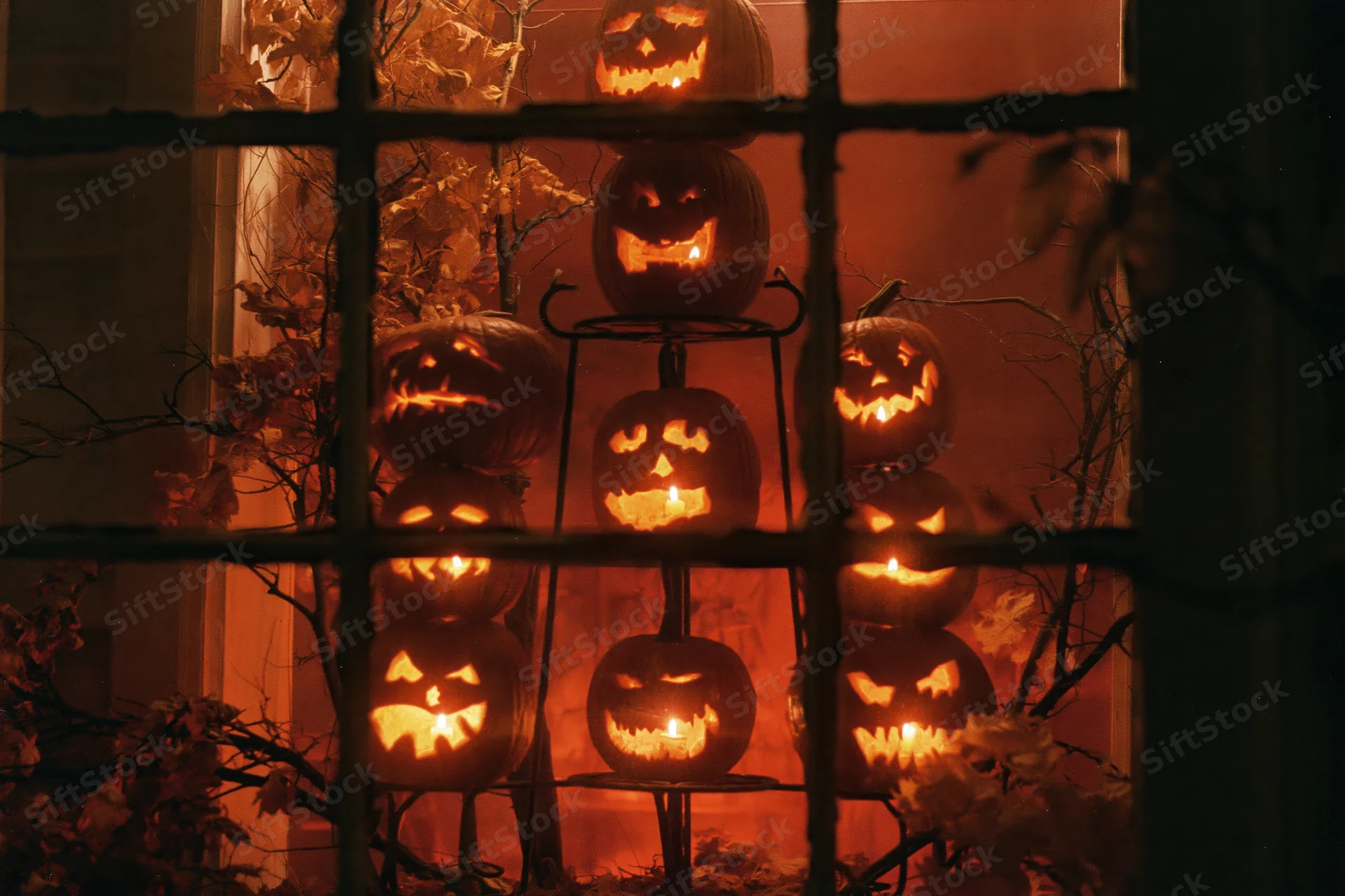 Seven glowing jack-o-lanterns stacked on a metal tiered stand viewed through a window grid at night