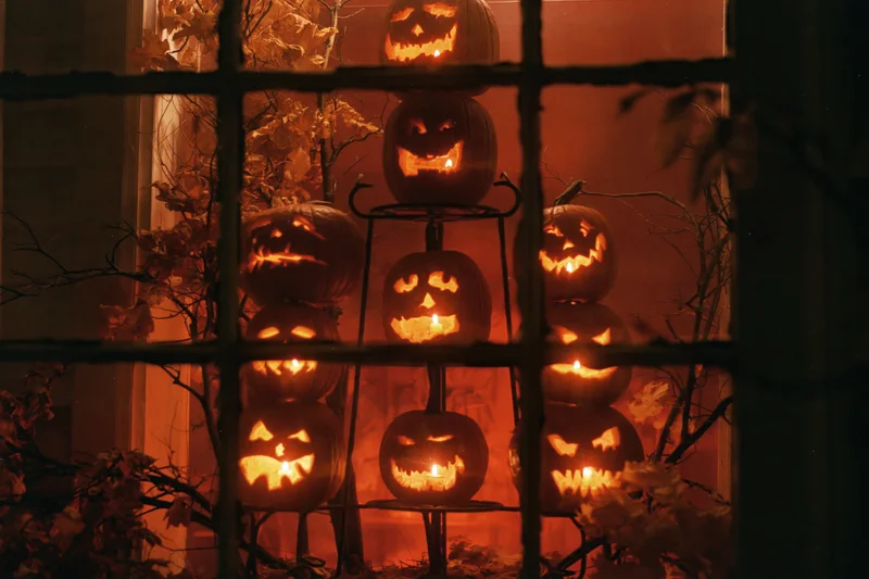 Seven glowing jack-o-lanterns stacked on a metal tiered stand viewed through a window grid at night