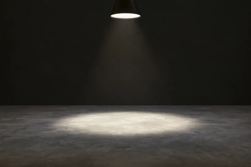 Single overhead spotlight casting a narrow cone of white light on a dark concrete floor in an empty room