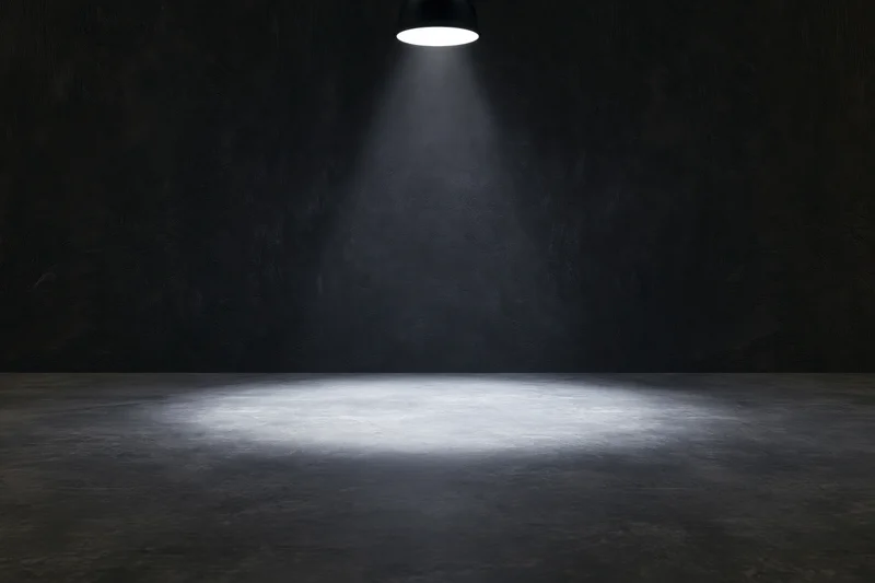Single overhead spotlight casting a narrow cone of white light on a dark concrete floor and wall in an empty industrial room