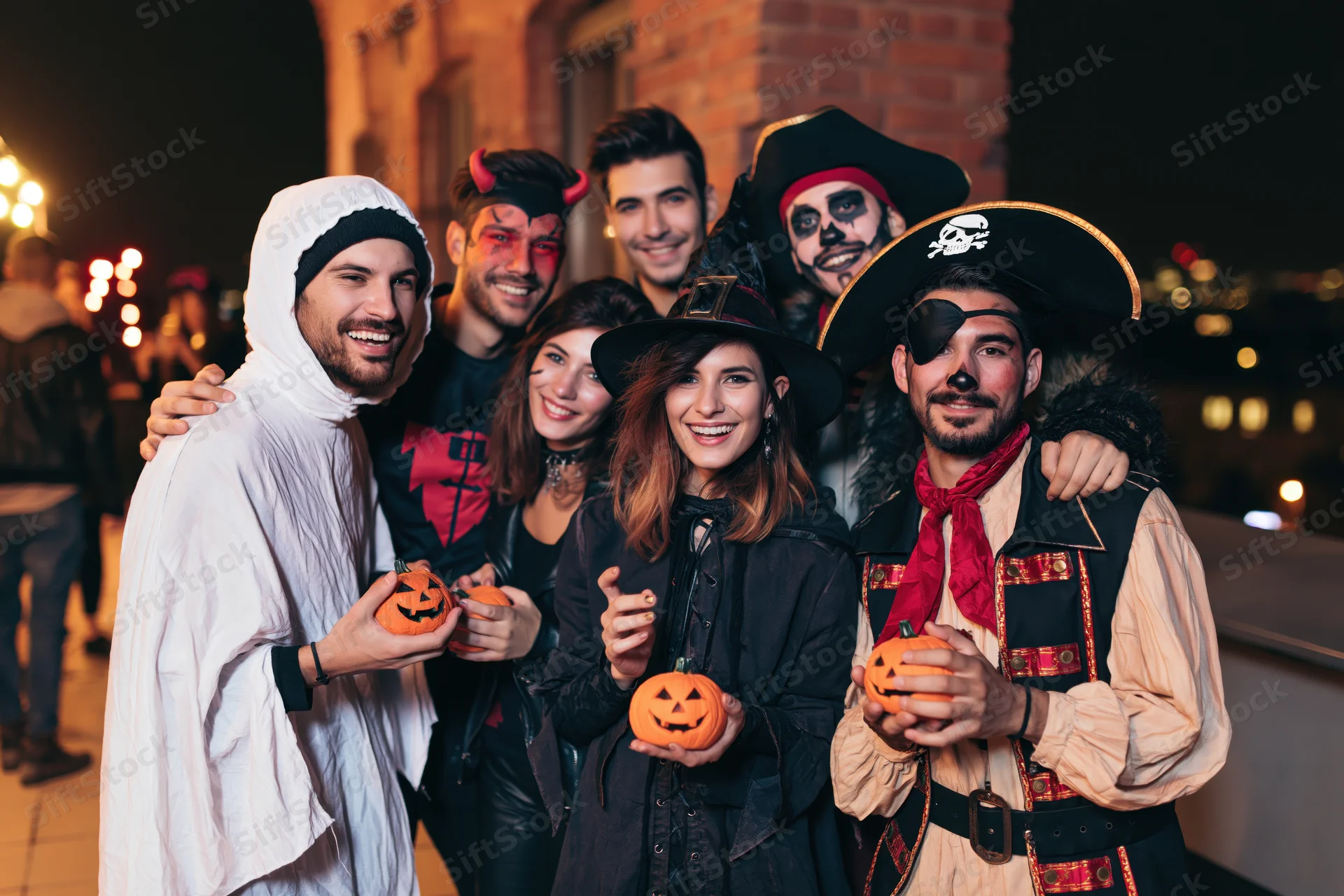 Six young adults in halloween costumes posing together outdoors at night holding pumpkins