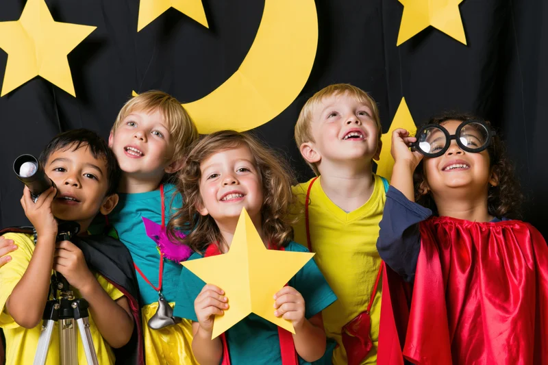 Five children in colorful costumes smiling and holding space-themed props against a black backdrop with yellow stars and moon