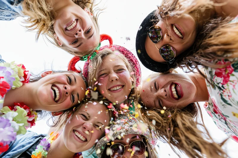 Six young women smiling down at camera in a circle with confetti falling around them outdoors