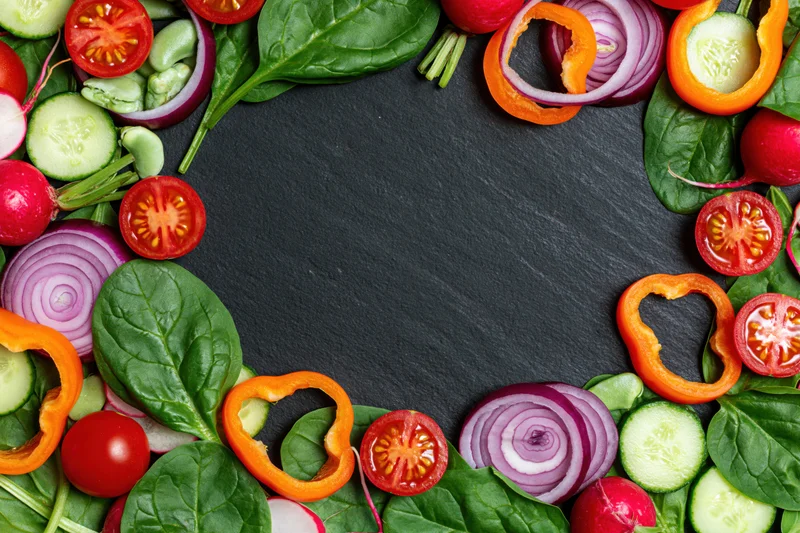 Fresh vegetables arranged in a circular border on dark slate surface with central copyspace