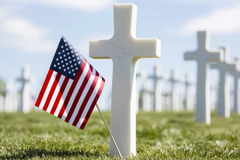 Small american flag planted at base of white marble cross in cemetery grass field