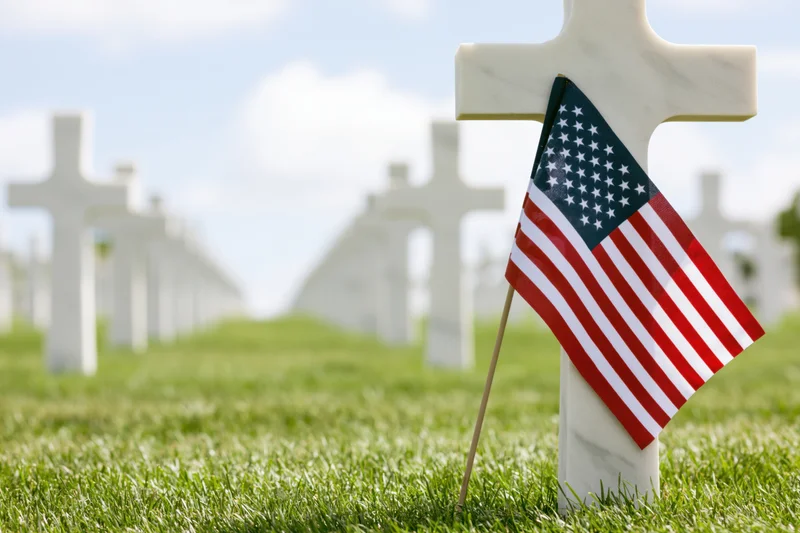 Small american flag planted at base of white marble cross in cemetery grass field