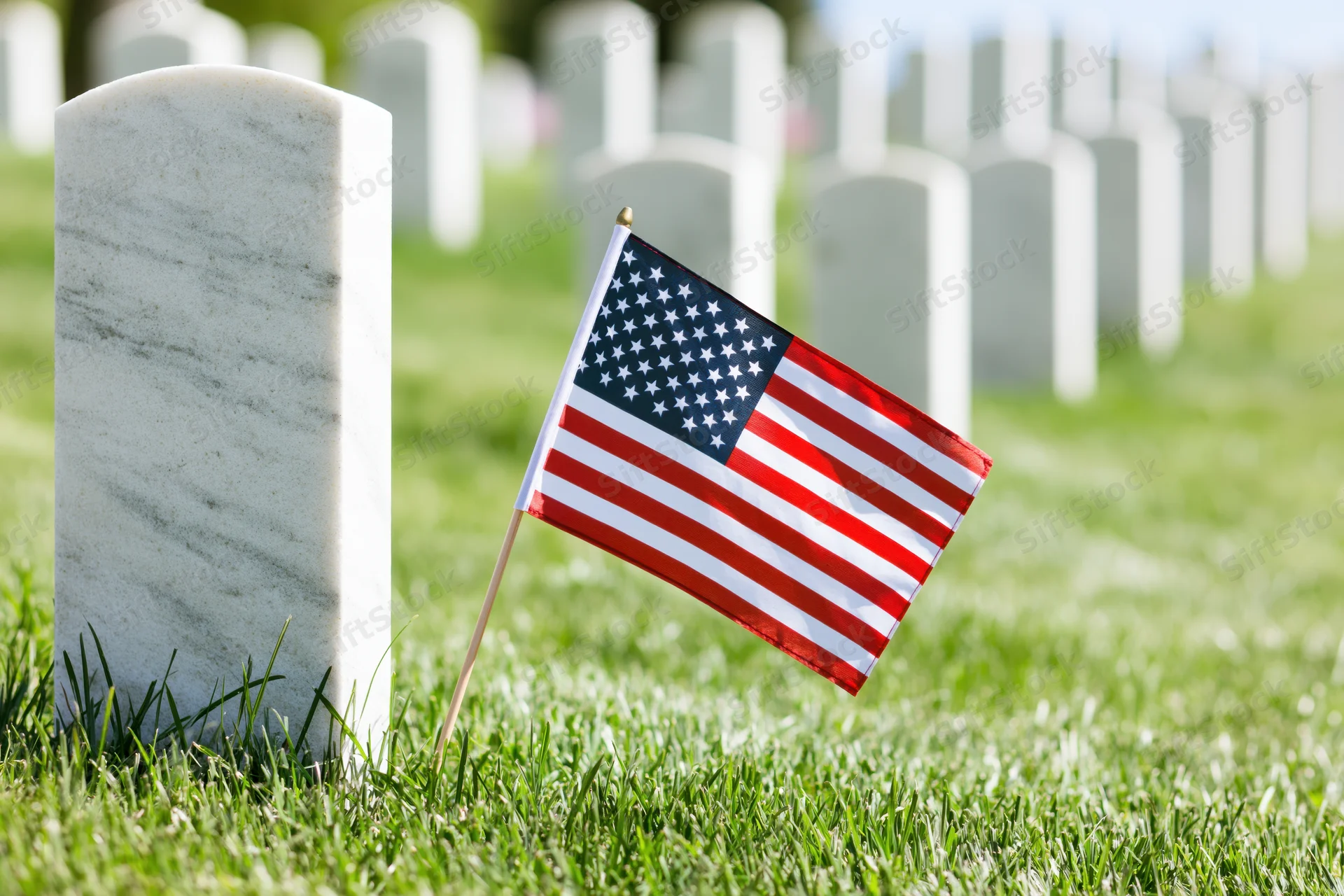 Small american flag planted in grass at base of white marble grave marker in cemetery