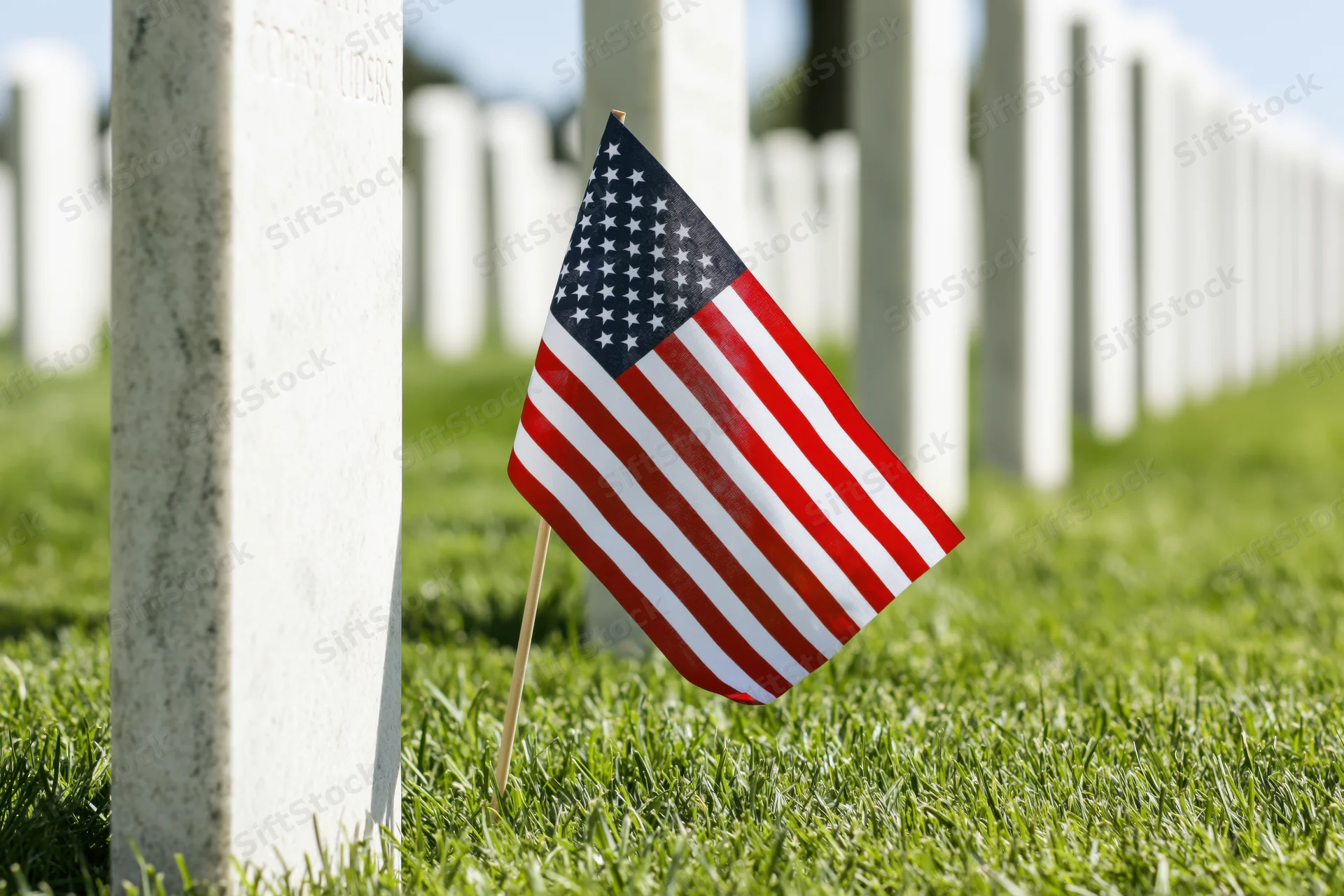 Small american flag planted in green grass at base of white tombstone in cemetery