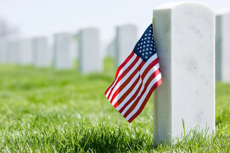 Small american flag placed at base of white tombstone in grassy cemetery