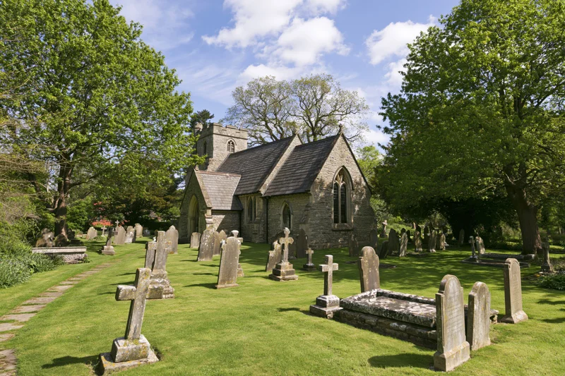Small victorian-era english parish church with stone walls and slate roof surrounded by graveyard and mature trees on sunny day