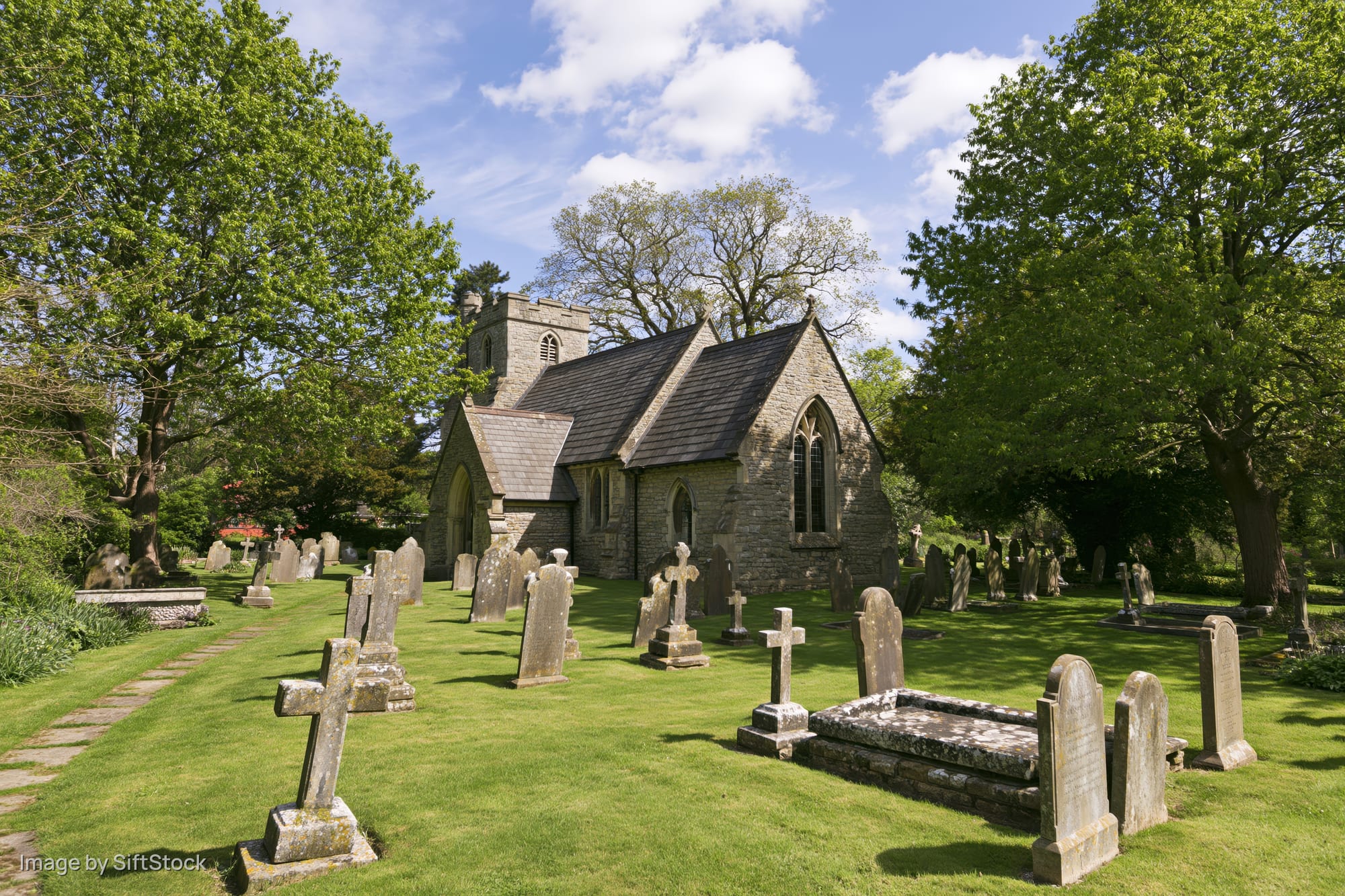 Small victorian-era english parish church with stone walls and slate roof surrounded by graveyard and mature trees on sunny day