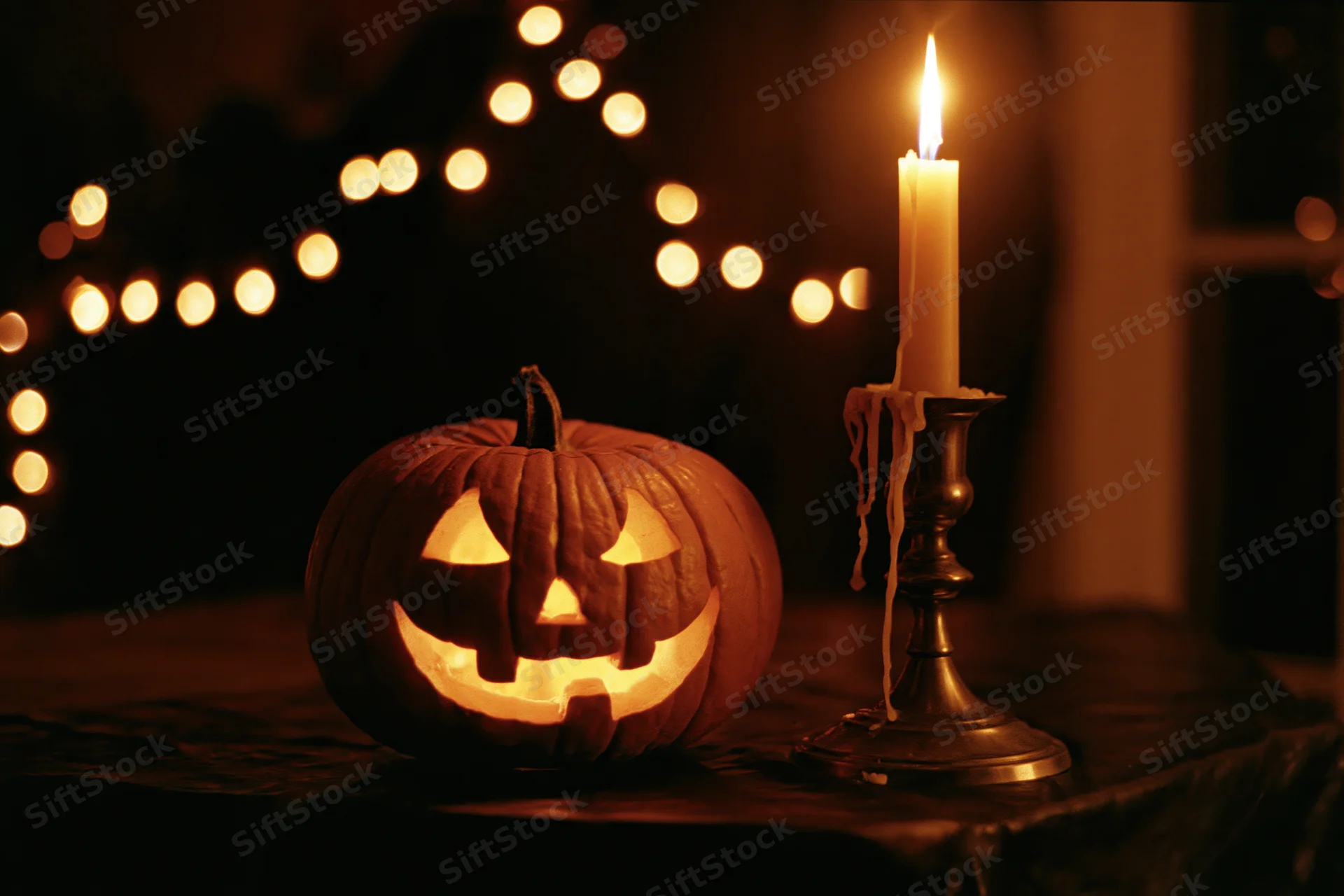 Carved jack-o-lantern glowing beside a lit candle on a dark table with string lights in the background