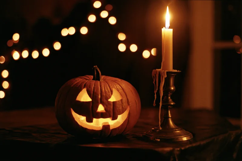 Carved jack-o-lantern glowing beside a lit candle on a dark table with string lights in the background