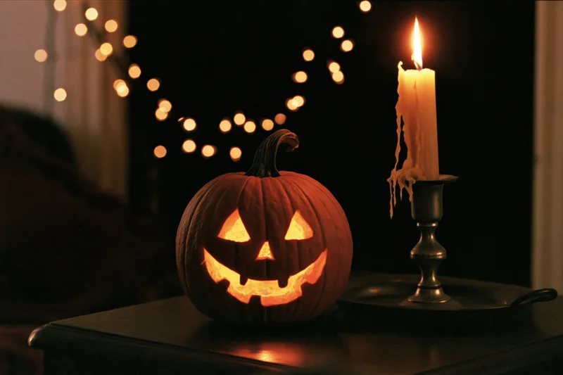 Carved jack-o-lantern glowing beside a lit candle on a dark table with string lights in the background