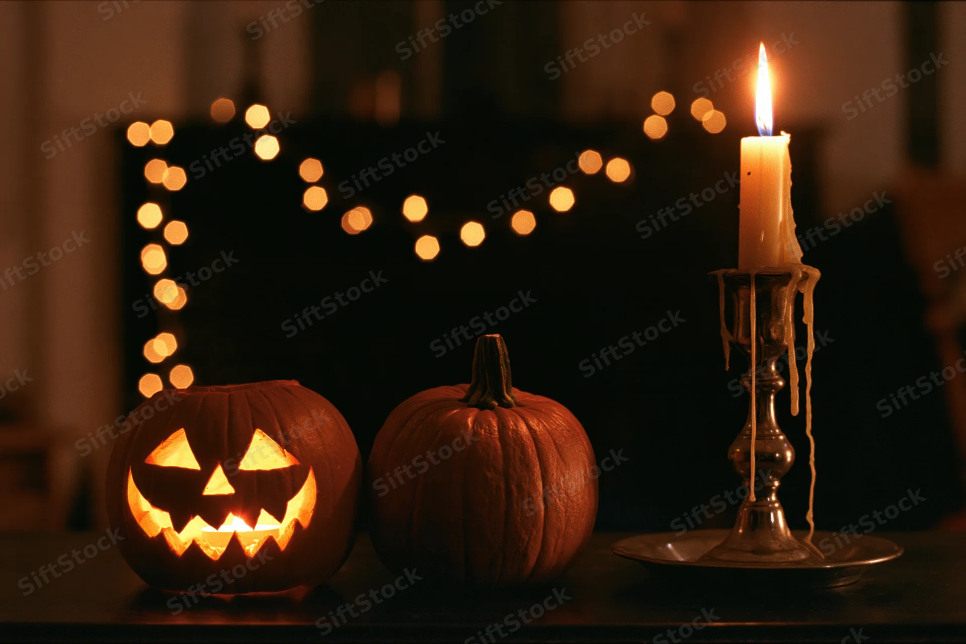 Carved jack-o-lantern glowing beside a whole pumpkin and lit candle on a dark table with string lights in the background