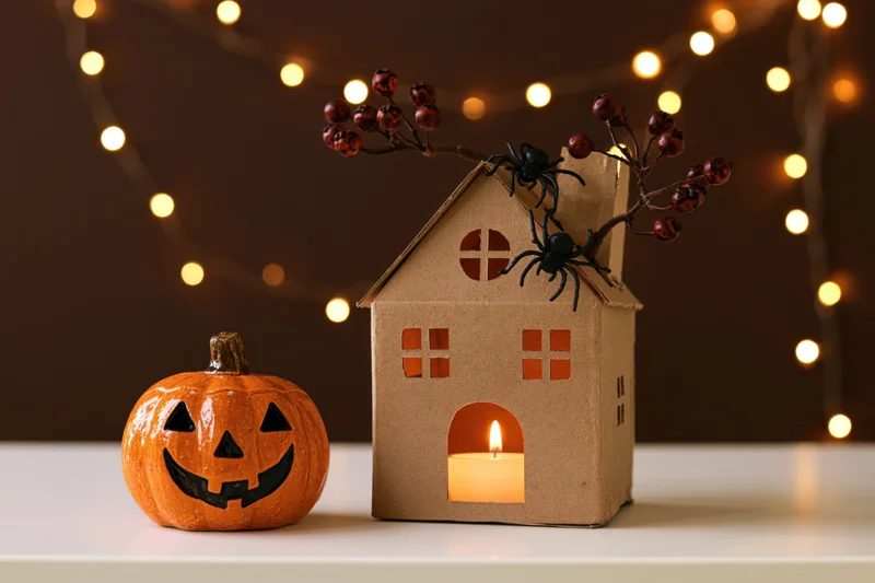 Small ceramic jack-o-lantern beside a handmade cardboard house with candle and spiders on tabletop
