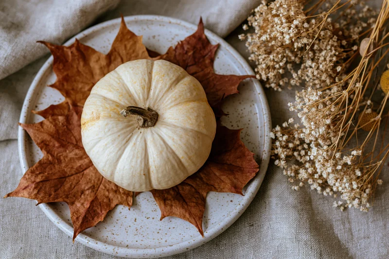 Small cream pumpkin on dried maple leaves arranged on a speckled ceramic plate with dried flowers beside it on linen fabric