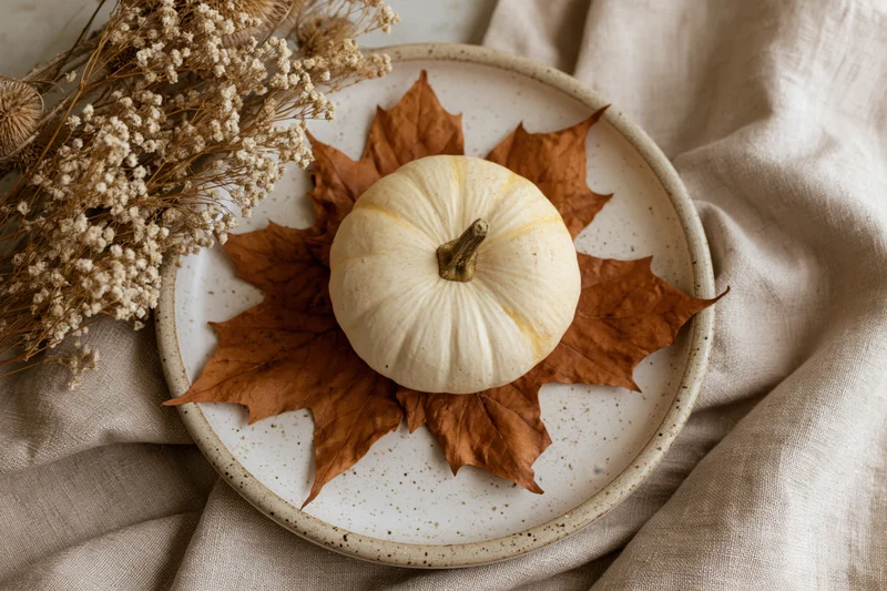 Small cream pumpkin on dried maple leaves arranged on a speckled ceramic plate with dried flowers on a linen cloth