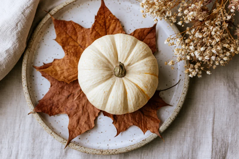 Small cream pumpkin on dried maple leaves and baby's breath on a speckled ceramic plate atop linen fabric
