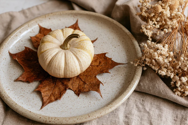 Small cream pumpkin resting on dried maple leaves on a speckled ceramic plate with dried flowers beside it